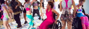 Photo by Kiam Marcelo Junio. A group of people wearing colorful, geometric Rebirth Garments dance during one of their performances. In the foreground, a dancer in a wheelchair is wearing a long, hot pink dress. The floor is painted with blue and yellow squares and shapes.