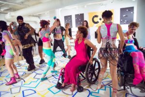 Photo by Kiam Marcelo Junio. A group of people wearing colorful, geometric Rebirth Garments dance during one of their performances. In the foreground, a dancer in a wheelchair is wearing a long, hot pink dress. The floor is painted with blue and yellow squares and shapes.