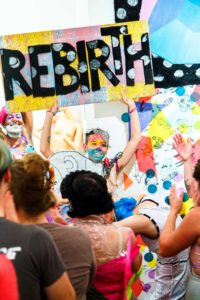 Photo by Grace Duval. Sky Cubacub, the founder of Rebirth Garments, holds up a large yellow-and-red checkerboard sign that says "Rebirth" in black letters. Sky has colorful, geometric makeup on and is surrounded by clapping, smiling people wearing colorful garments.